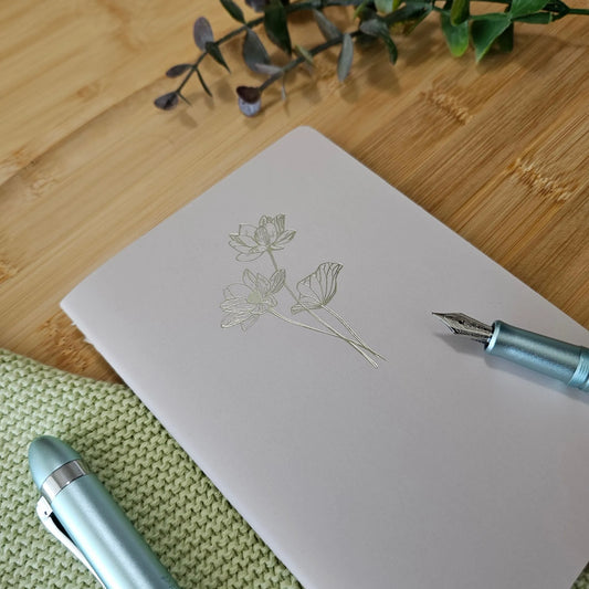 Artchemira A6 pocket journal in muted rosy with gold floral foil, blue pen on bamboo desk