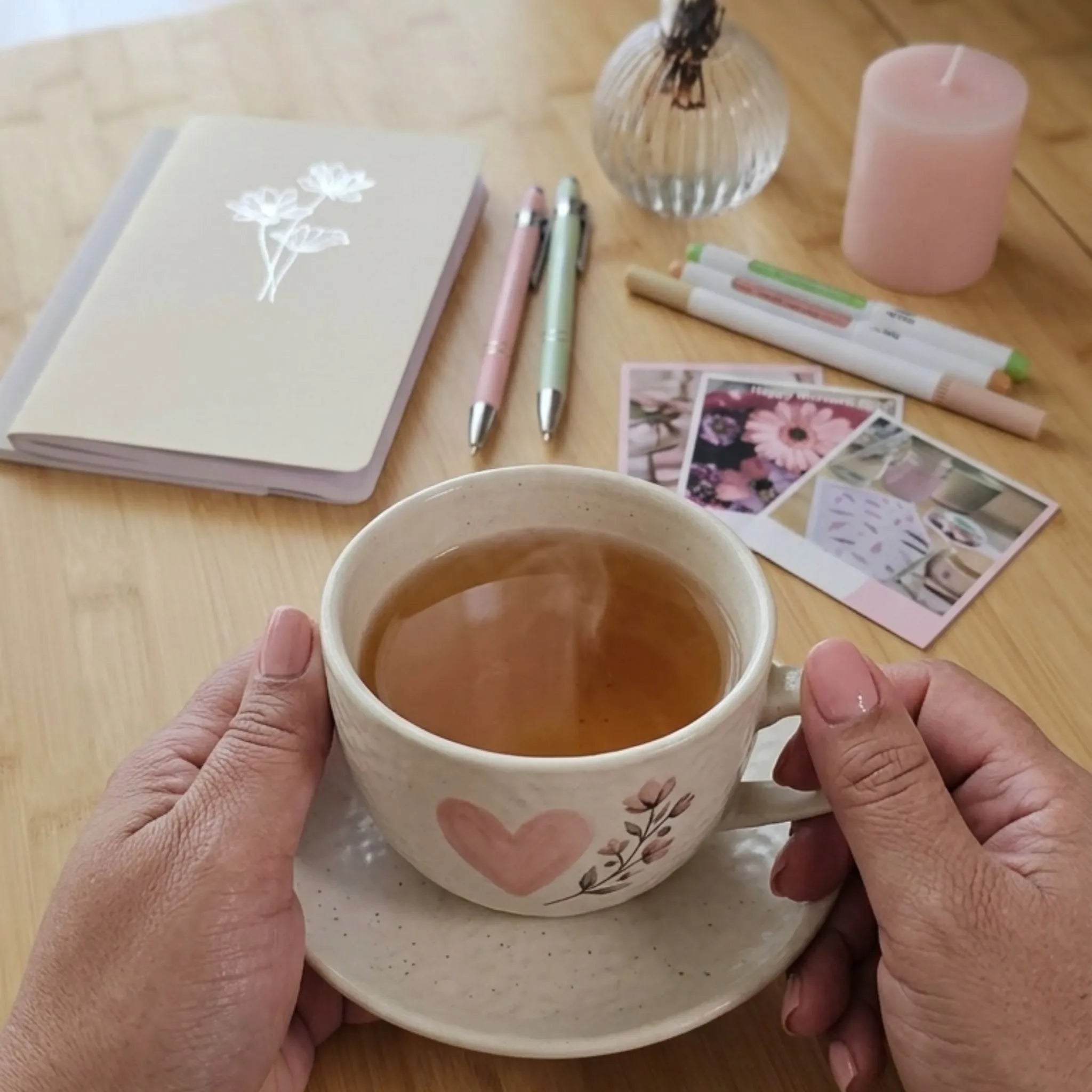 Hands cradling a heart-motif tea cup beside an Artchemira floral journal, pens, and photo cards on a wooden desk — evoking slow, mindful journaling