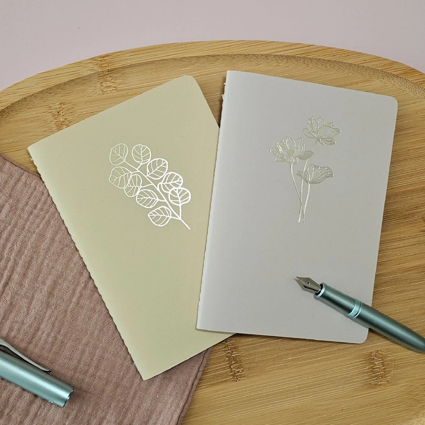Artchemira A6 pocket journals, in dusty rose and warm sand  with champagne gold foil covers featuring  Lotus and Eucalyptus design on bamboo flat lay