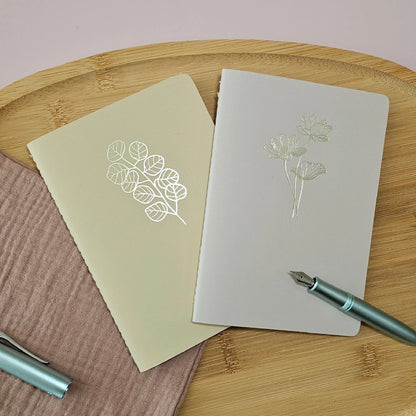 Artchemira A6 pocket journals, in dusty rose and warm sand  with champagne gold foil covers featuring  Lotus and Eucalyptus design on bamboo flat lay