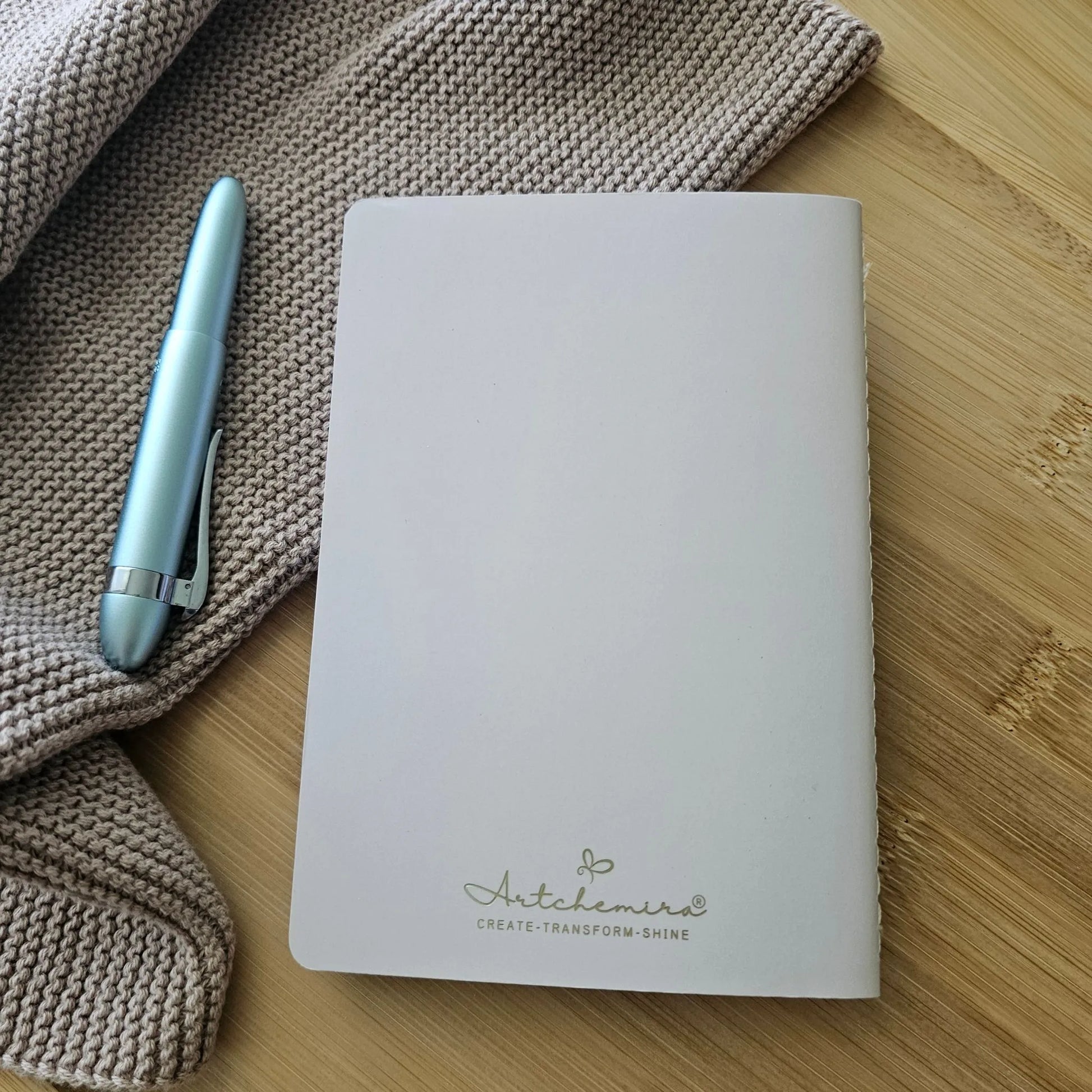 Artchemira A6 pocket journal back cover  in muted rose with champagne gold foil logo, blue pen on knit fabric and bamboo desk