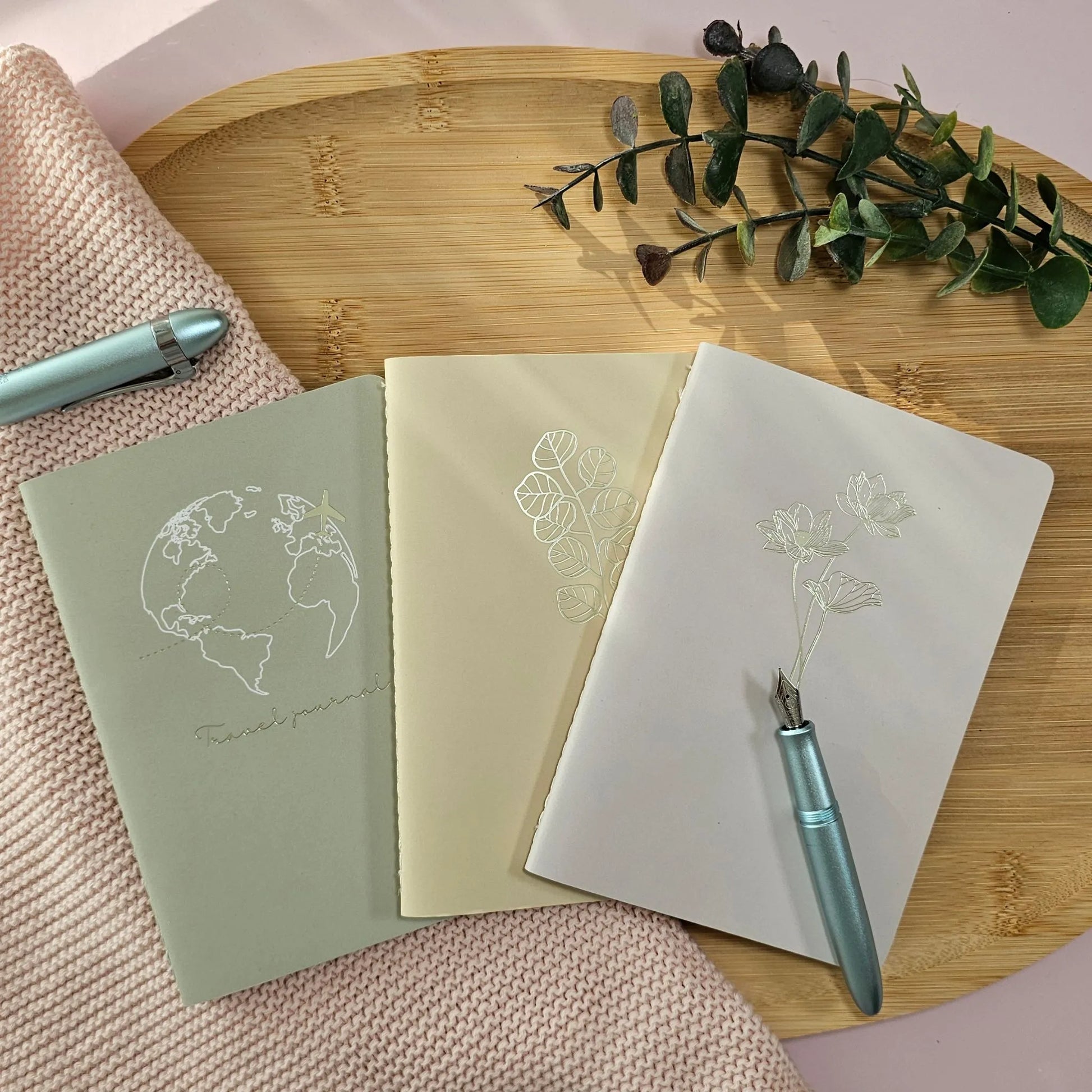Artchemira A6 pocket journal trio in sage,warm sand  and dusty rose tones with champagne gold foil covers featuring Globe and Plane, Eucaliptus and Lotus design on bamboo plate