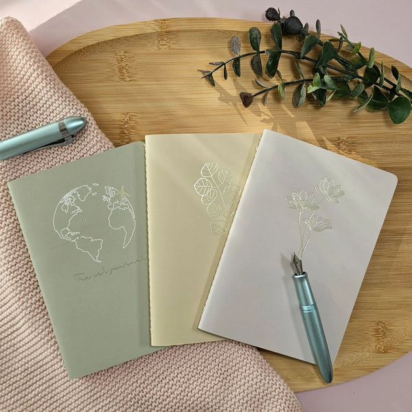 Artchemira A6 pocket journal trio in sage,warm sand  and dusty rose tones with champagne gold foil covers featuring Globe and Plane, Eucaliptus and Lotus design on bamboo plate