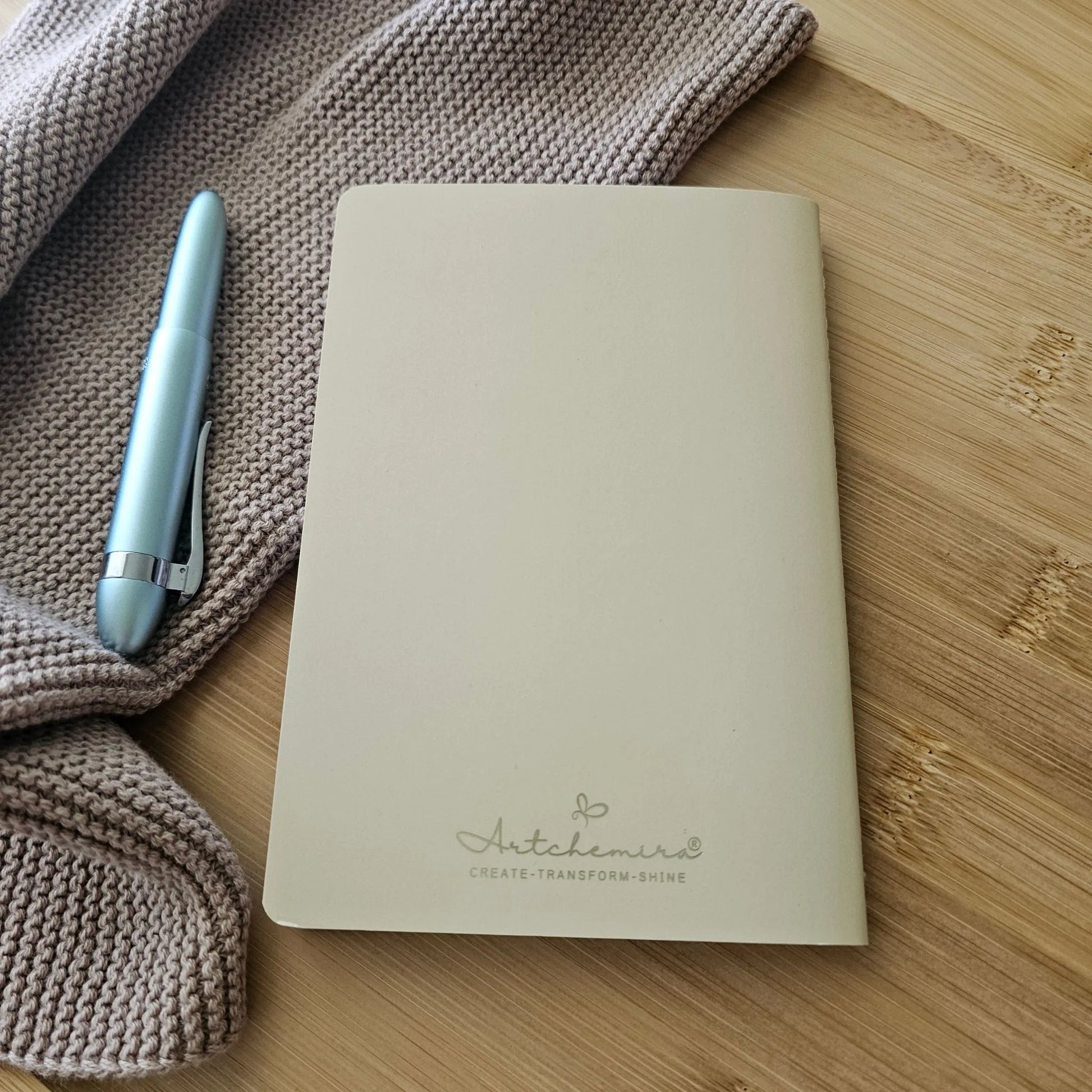Artchemira A6 pocket journal back cover in warm sand tone and champagne gold foil logo, on bamboo desk