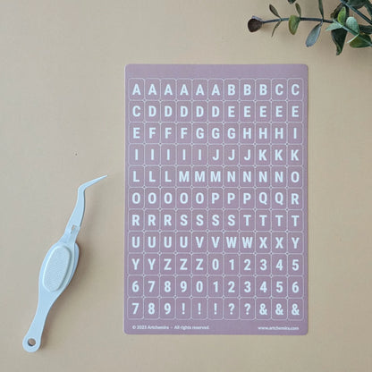 Artchemira dusty pink alphabet sticker sheet with white crafting tweezer tool
on beige surface