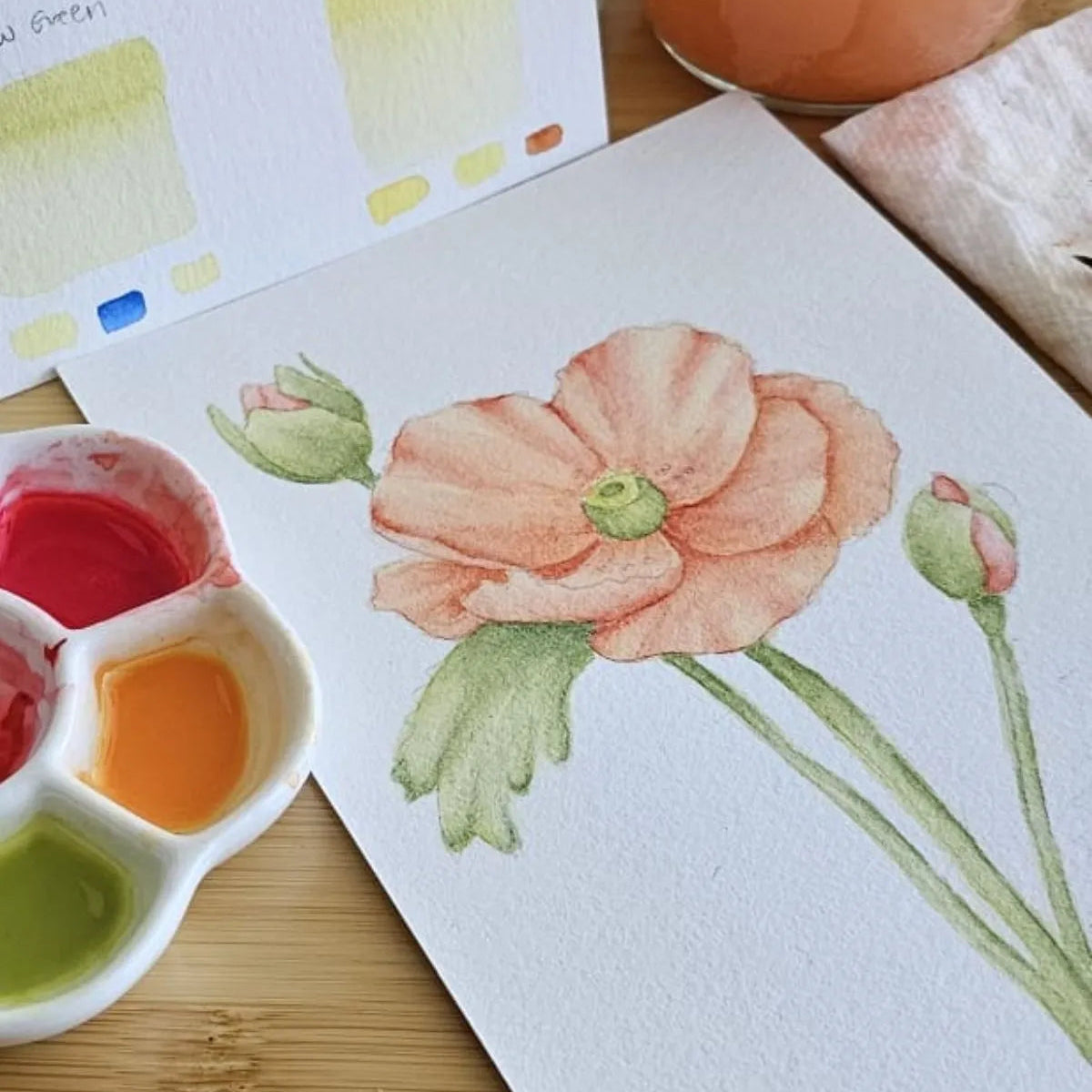 Watercolour painting of a peach poppy flower with buds, beside a ceramic palette with vibrant pigments and colour swatches — Artchemira watercolour practice