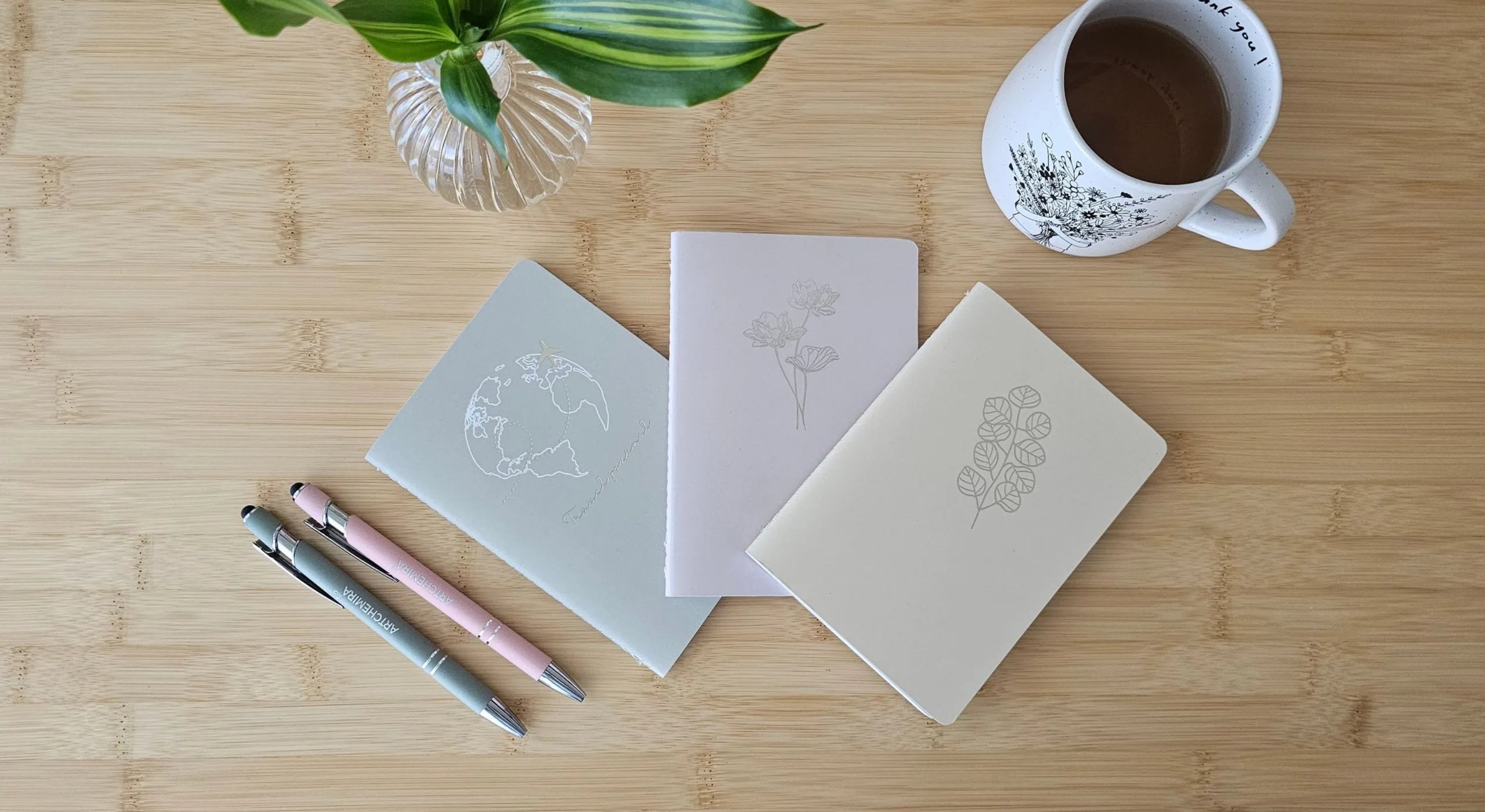 Flat lay of minimalist notebooks, pen, coffee cup, and flowers on wooden table — representing calm morning journaling and mindful creativity