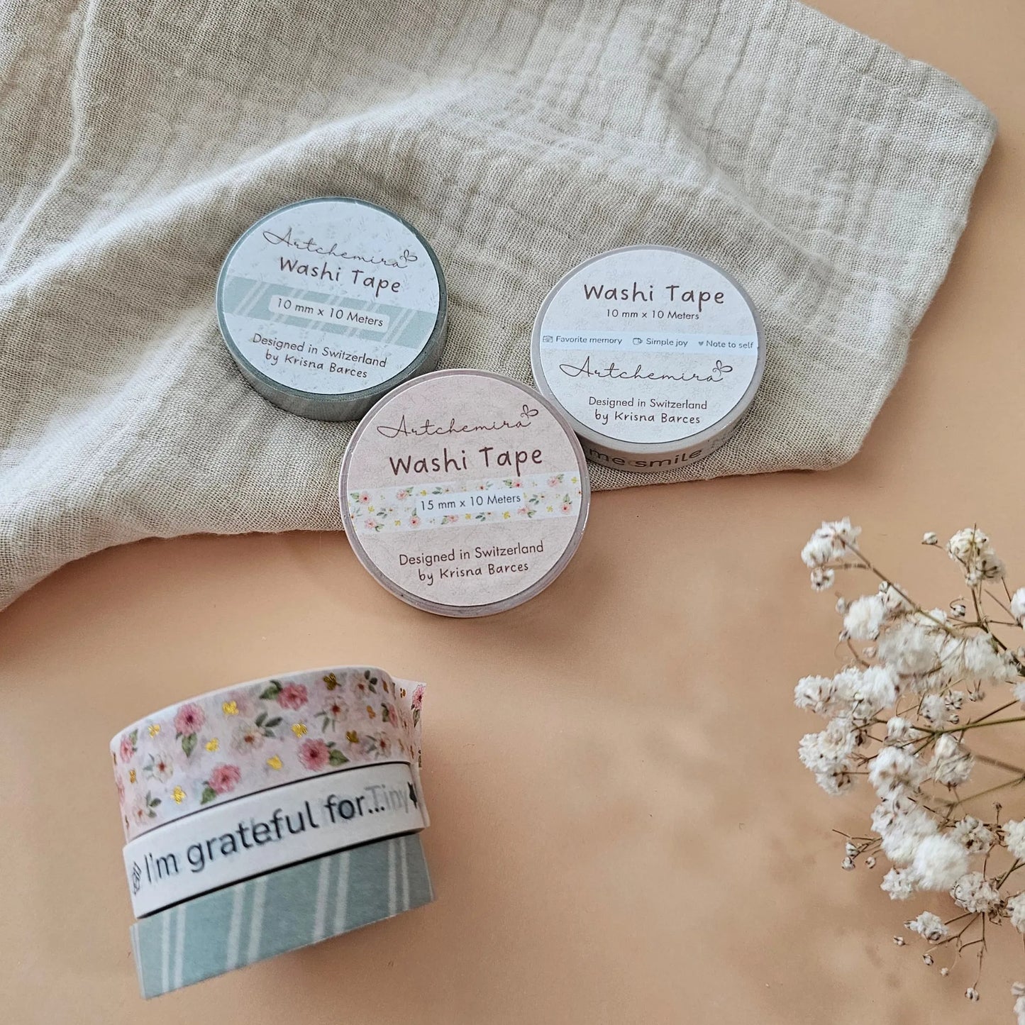 Three Artchemira rolls of washi tape on a beige surface with a light fabric and flowers in the background.